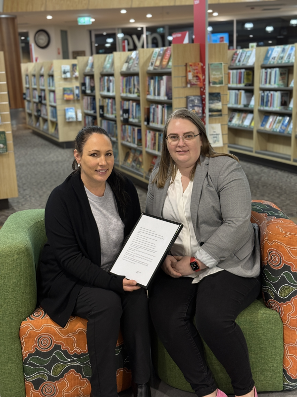 Community Development Coordinator Kristi Brooks (left) and Acting Manager Library and Community Development Sally Mann with one of the framed stories “I grew up quickly. My childhood disappeared into the bush, running, hiding surviving …”